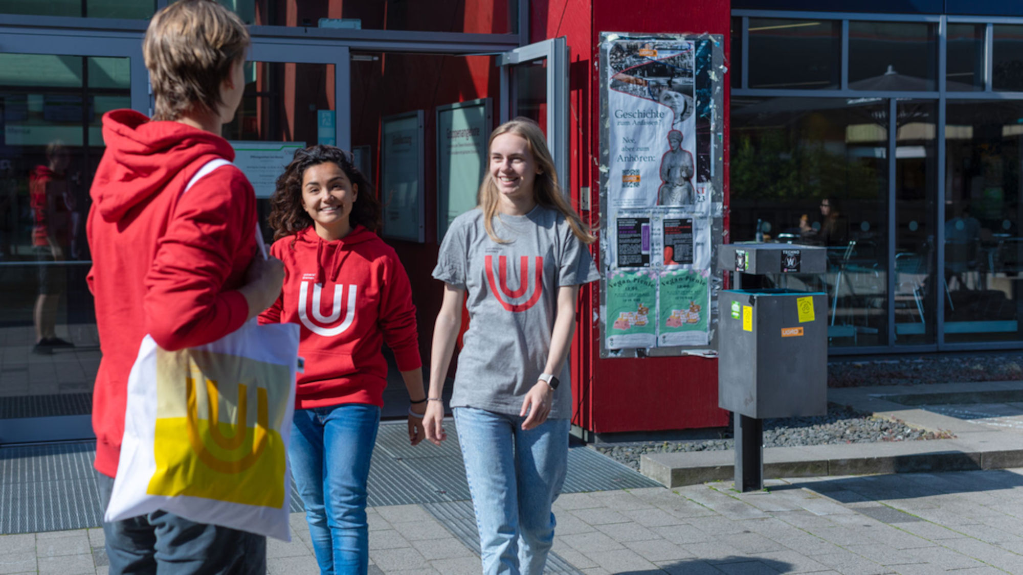 A young man stands in front of the Mensa cafeteria, a cotton bag with the University of Bremen logo hanging over his shoulder. He greets two female students coming out of the Mensa cafeteria.