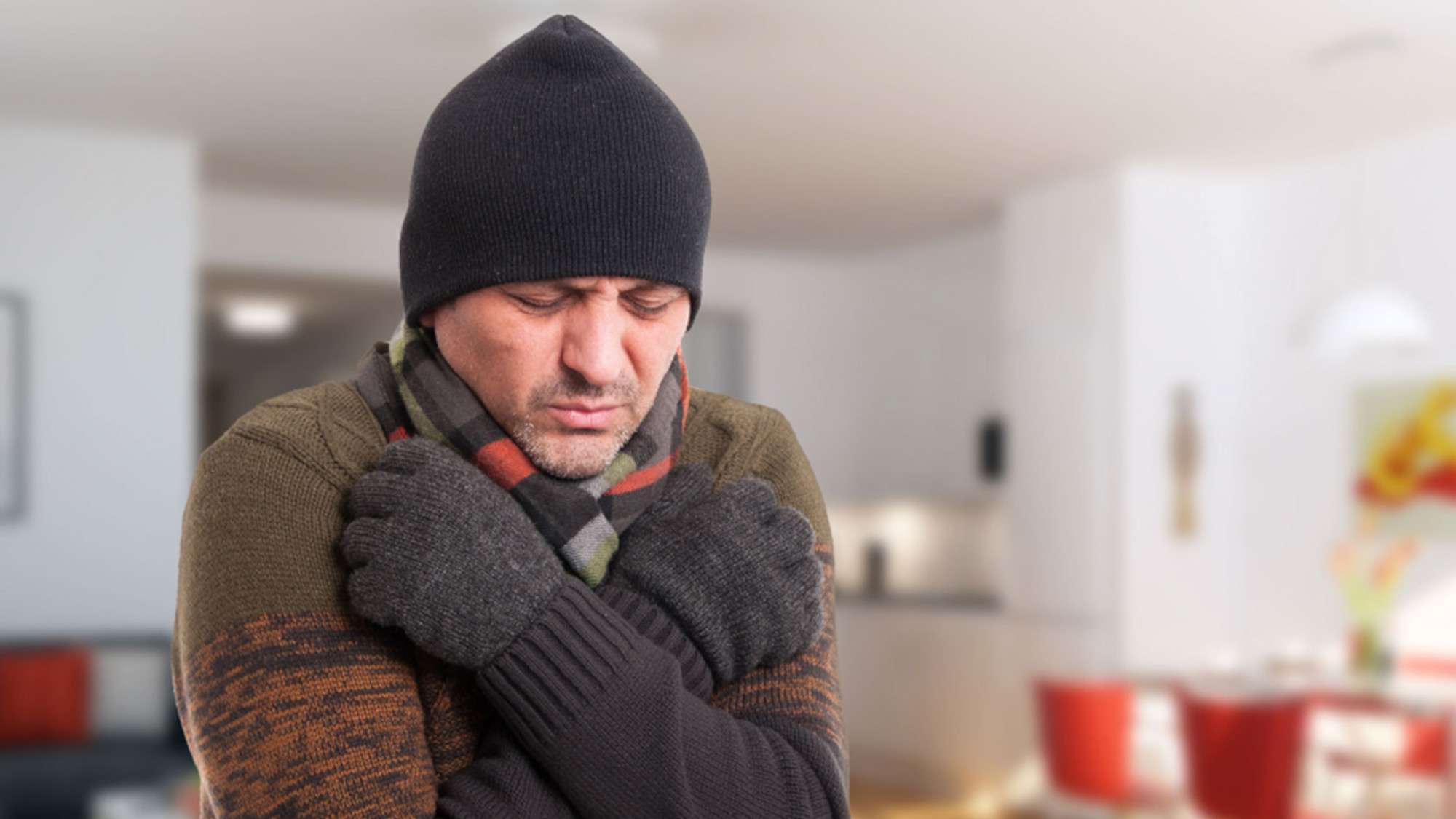 Ein Mann mit Schal und Mütze steht im Wohnzimmer und umarmt sich frierend selbst.