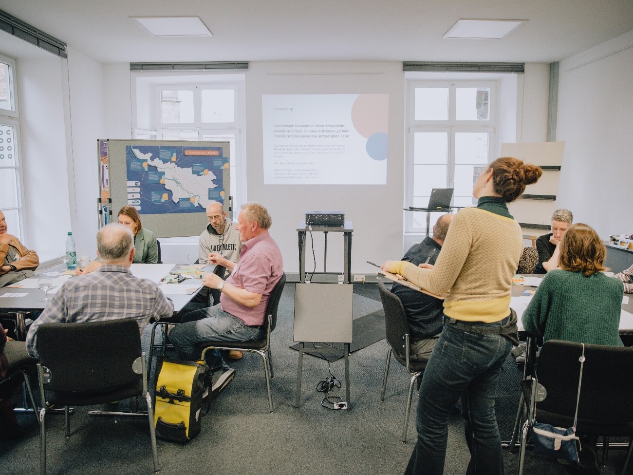 The photo shows a room with two large group tables where adults are seated. In the background, there is a map and a presentation projected onto the screen. Standing in front of them in the room is a person holding a clipboard. 