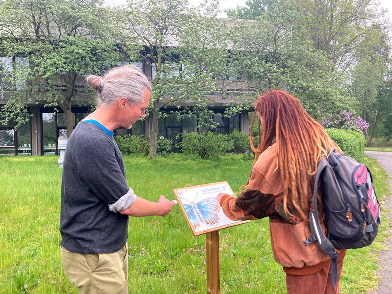 Marko Rohlfs und Lorena Kalvelage stehen vor einer ihrer Informationstafeln neben einer Wiese, auf der ein großer blauer Schmetterling zu sehen ist.