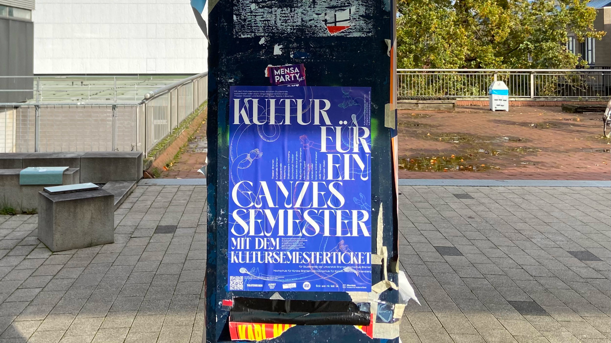 An einem Pfeiler auf dem Universitäts-Boulevard hängt ein blaues Plakat, welches über das Kultursemesterticket informiert.