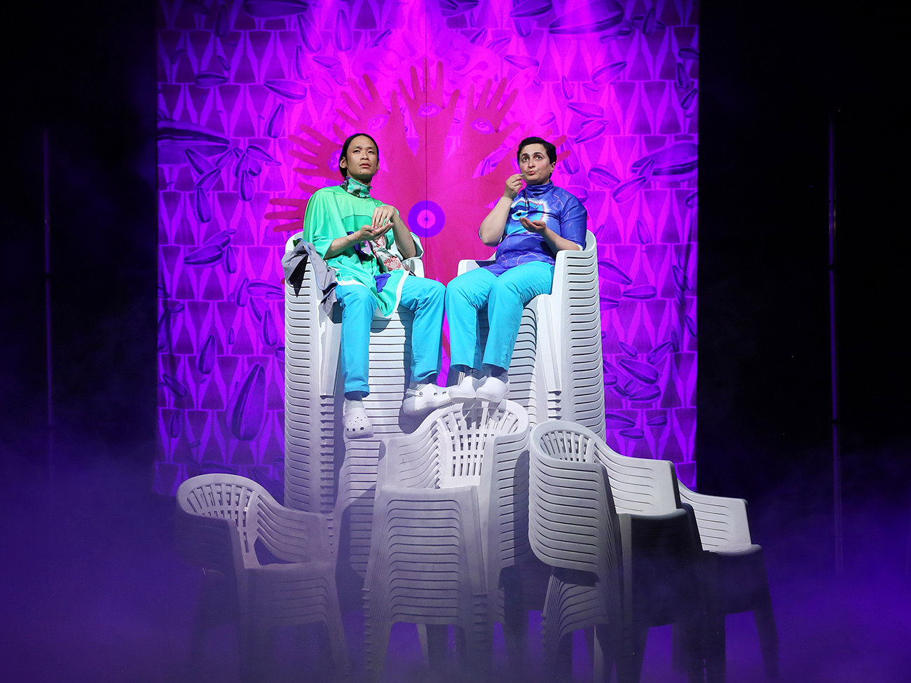 Auf der Bühne sitzen zwei Personen auf einem hohen Stapel an Plastikstühlen. Hinter ihnen ist ein grell-lilaner Vorhang zu sehen.