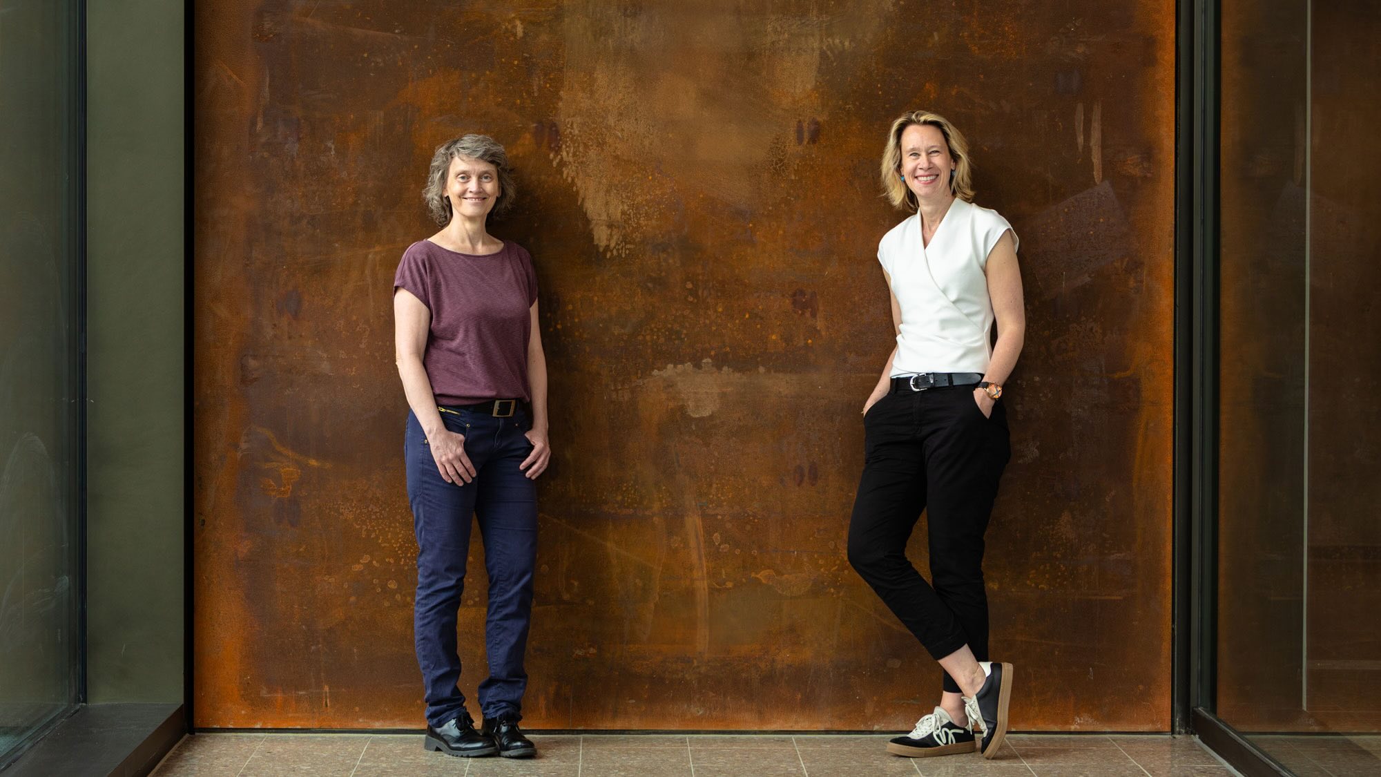 Die Professorinnen Rita Groß-Hardt und Karen Struve lehnen gegen eine braune Wand und lächeln in die Kamera.