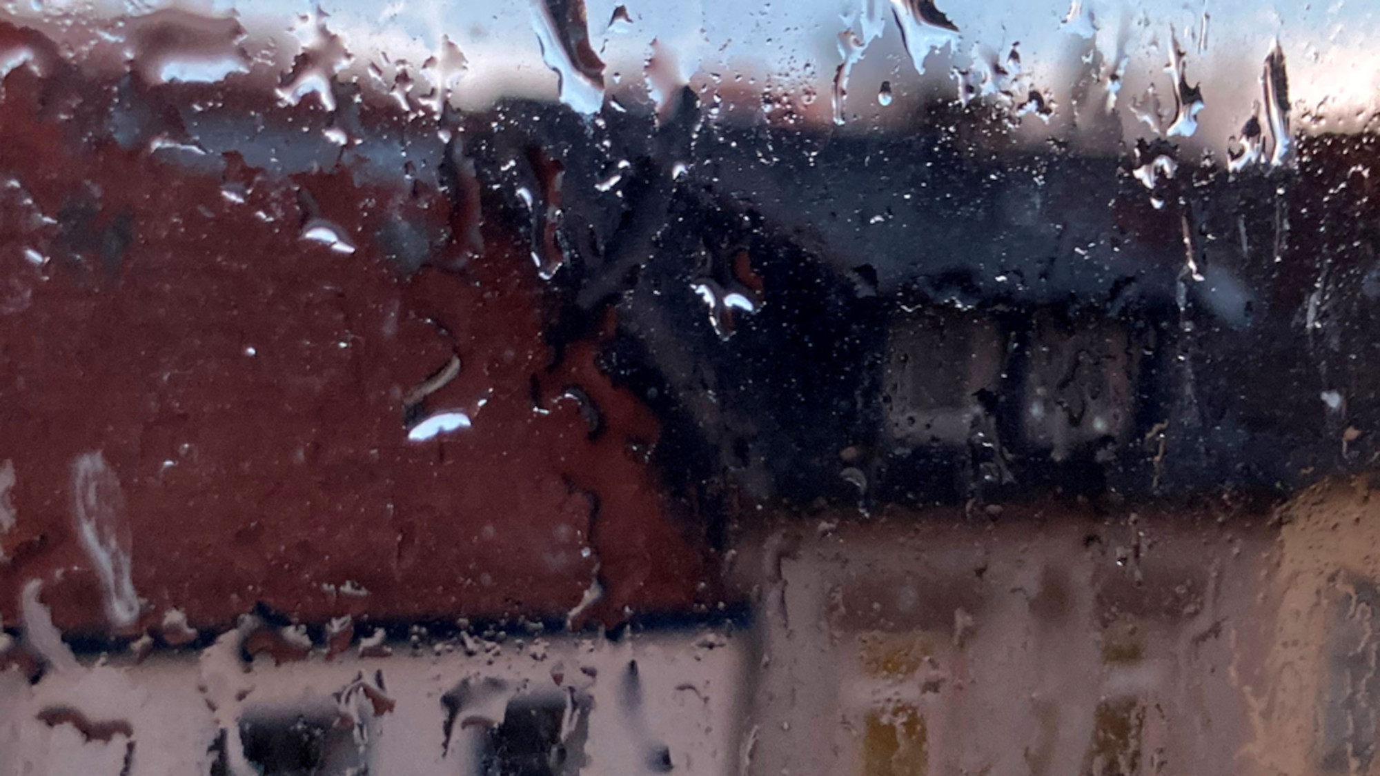 Regen an der Fensterscheibe, im Hintergrund sind verschwommen Häuser zu erkennen.
