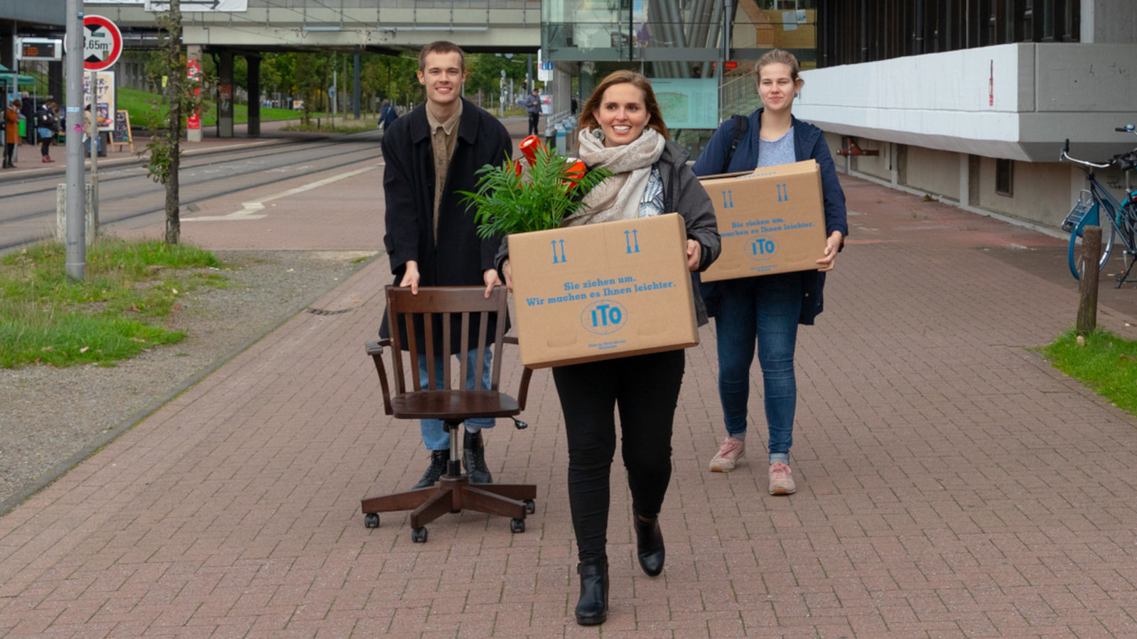 Drei junge Personen laufen lächelnd und mit Umzugskartons über den Campus der Uni Bremen