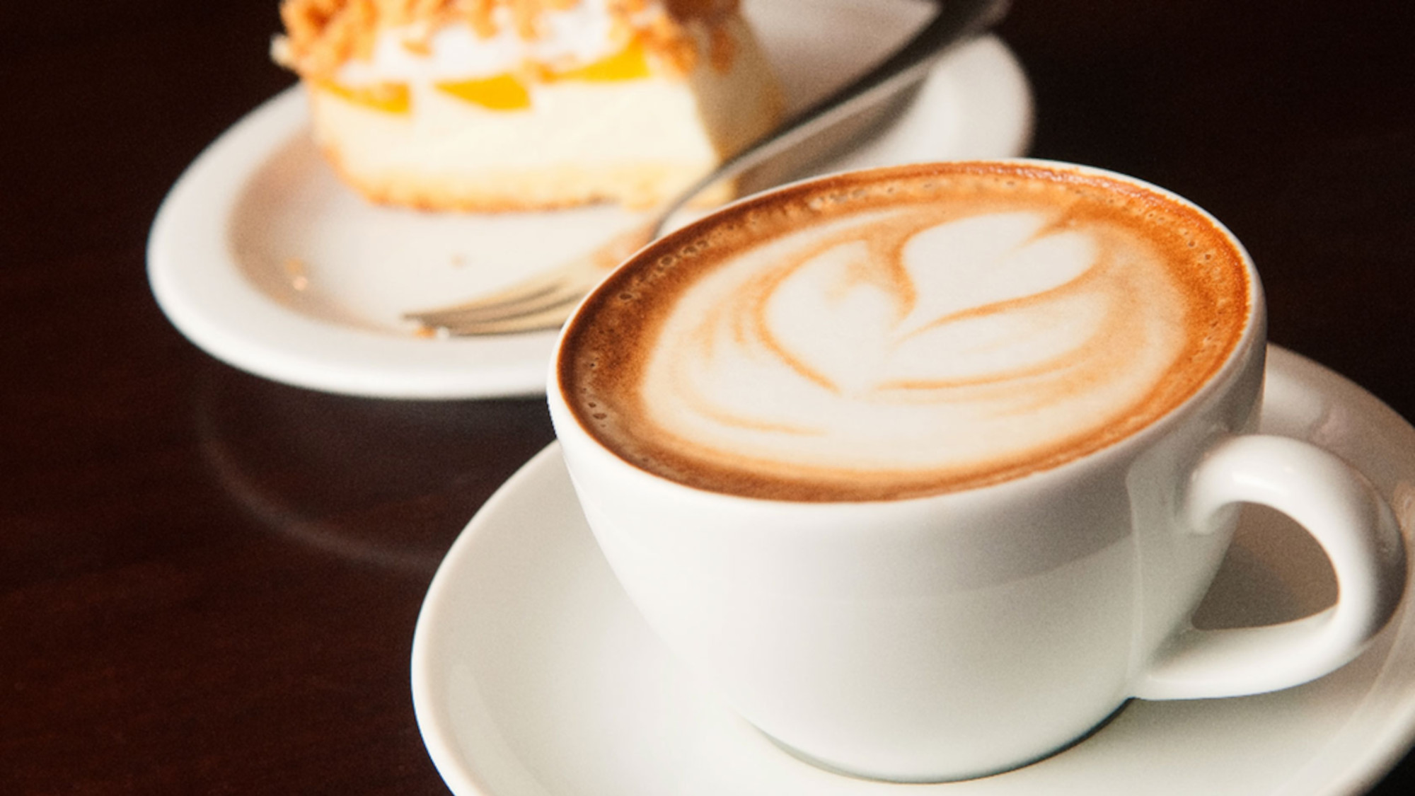 Eine Cappuccino-Tasse steht auf einem Tisch, im Hintergrund ist ein Stück Apfelkuchen zu sehen.