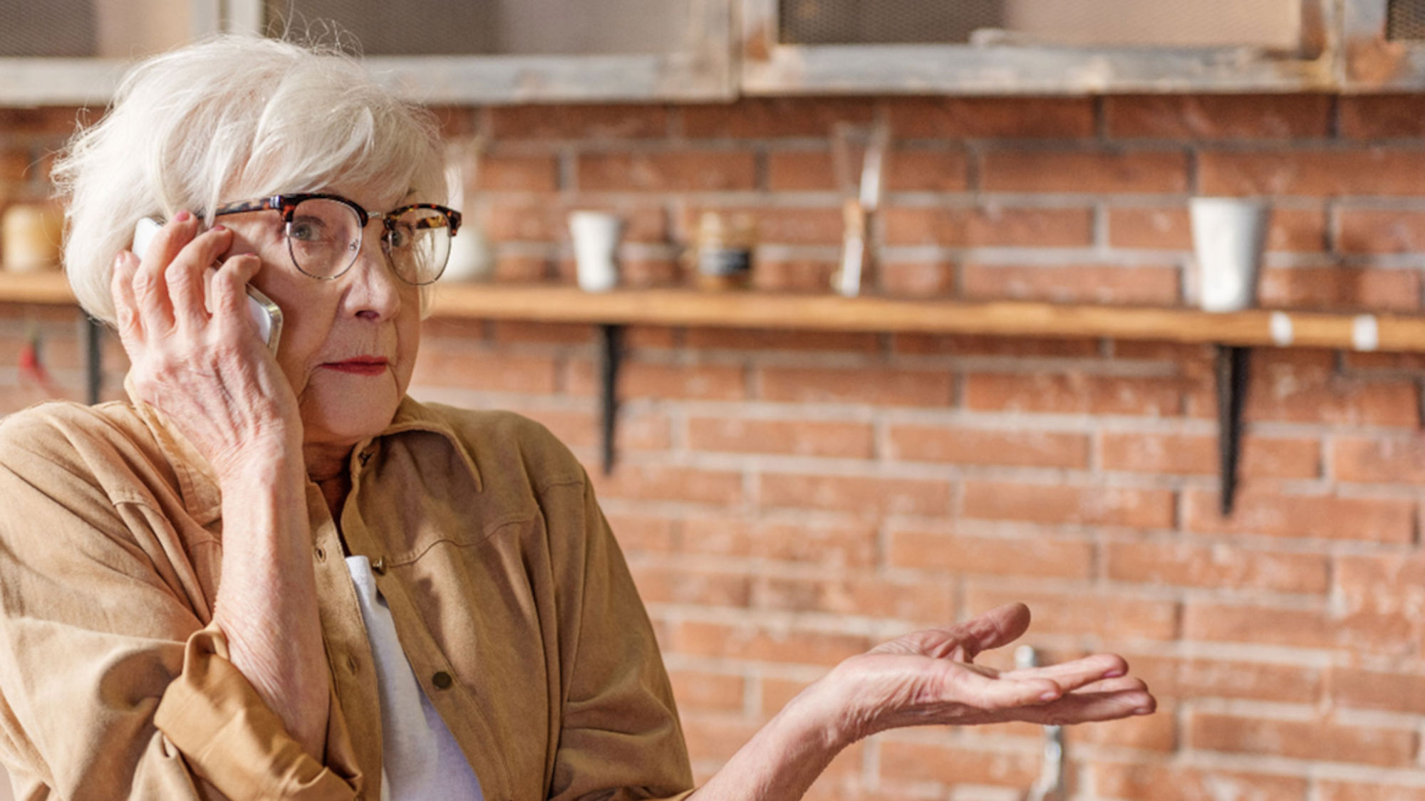 An elderly woman holds her cell phone to her ear, looking confused